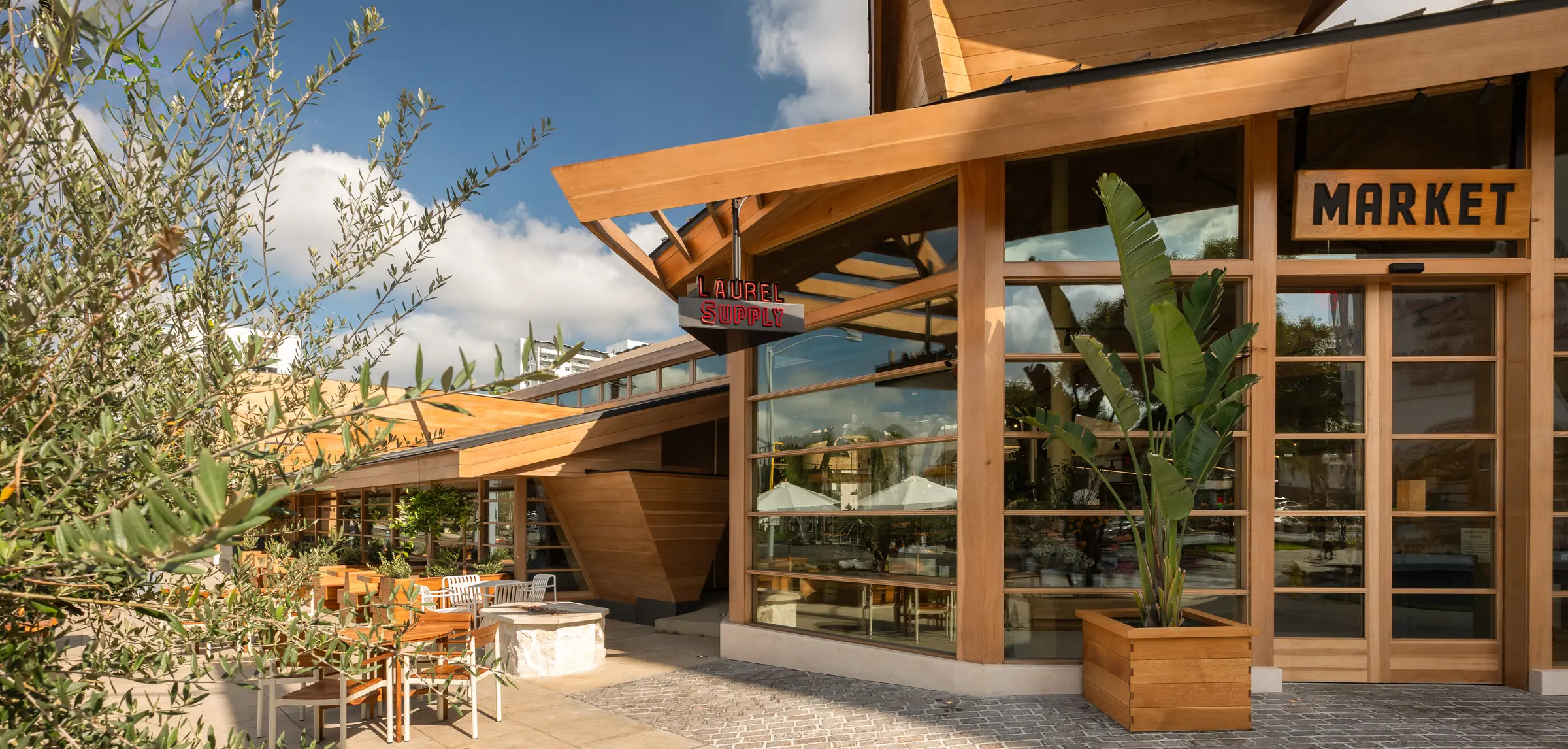 The Laurel Supply market entrance under its timber roof, with olive trees and patio seating